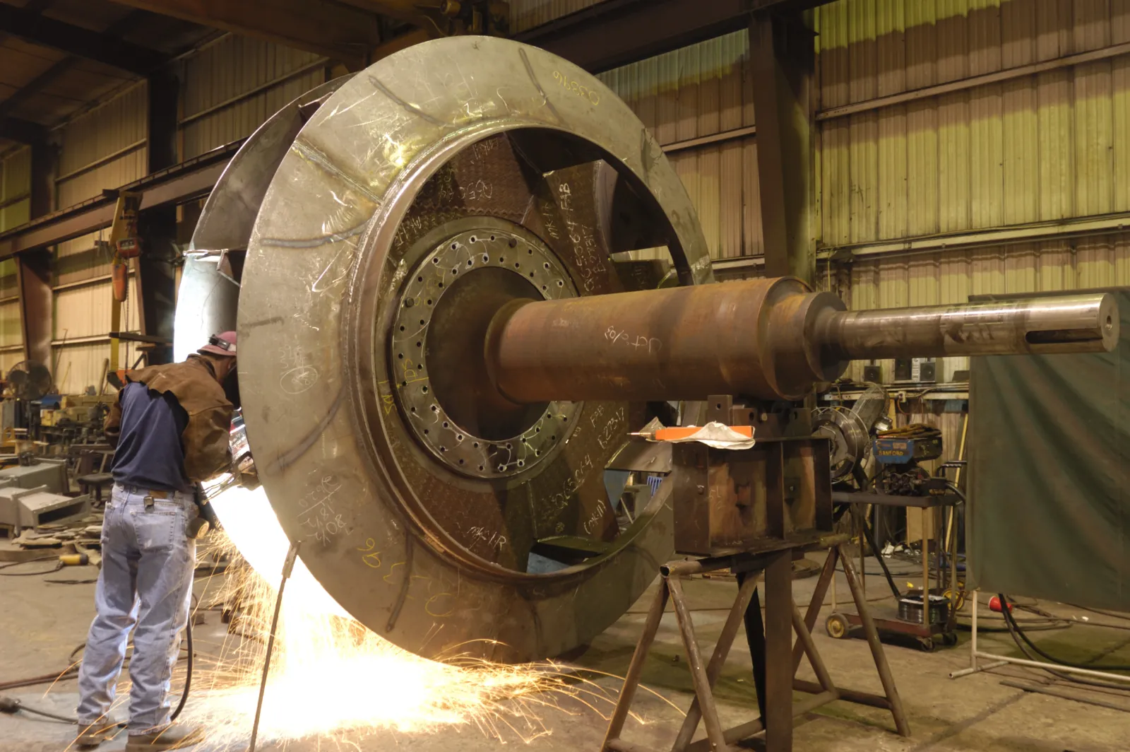 Welder repairing a large centrifugal fan wheel on a stand, sparks flying, in the Barron Fan shop